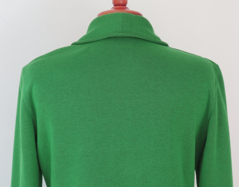 May include: A bright green knit cardigan featuring a shawl collar and long sleeves. The garment is constructed from a solid-coloured, textured knit fabric and is displayed on a red mannequin neck piece, showing the back view of the upper torso.