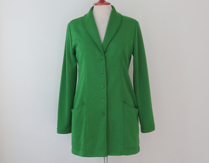 May include: A vibrant grass green blazer featuring a shawl collar, long sleeves, and a front button closure with five matching green buttons. The garment includes two patch pockets on the lower front and vertical seam detailing along the torso.