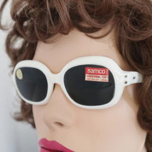 Może przedstawiać: Białe owalne okulary przeciwsłoneczne z ciemnymi soczewkami. Okulary przeciwsłoneczne mają naklejkę z napisem "samco" i "ASSORB. 75% CONTROLLED LENSES". Zausznik okularów jest ozdobiony małymi detalami w kształcie gwiazd.