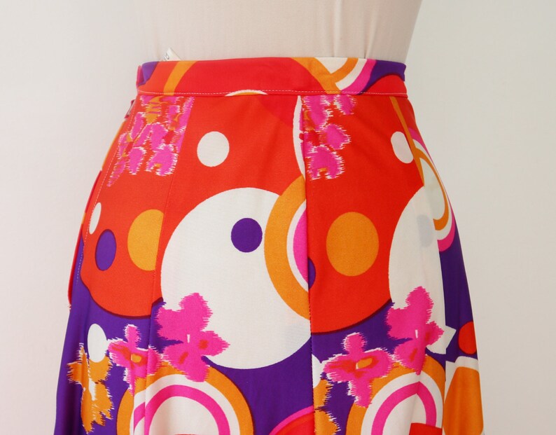 May include: A vibrant, high-waisted skirt featuring a bold, retro design. The skirt showcases a colorful pattern of large circles, flowers, and geometric shapes in shades of orange, white, purple, and pink. The fabric appears to be lightweight and flowing.