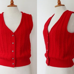 Puede incluir: Un chaleco sin mangas de color rojo brillante con cuello en V y diseño de punto trenzado. El chaleco tiene un dobladillo acanalado y se abrocha con cinco botones plateados. Se muestra en un maniquí.