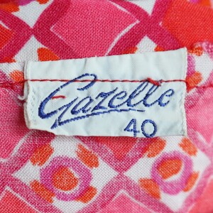 May include: Close-up of a fabric with a repeating pattern of pink, red, and white geometric shapes. A white label with the blue text "Gazelle" and the number "40" is sewn onto the fabric.