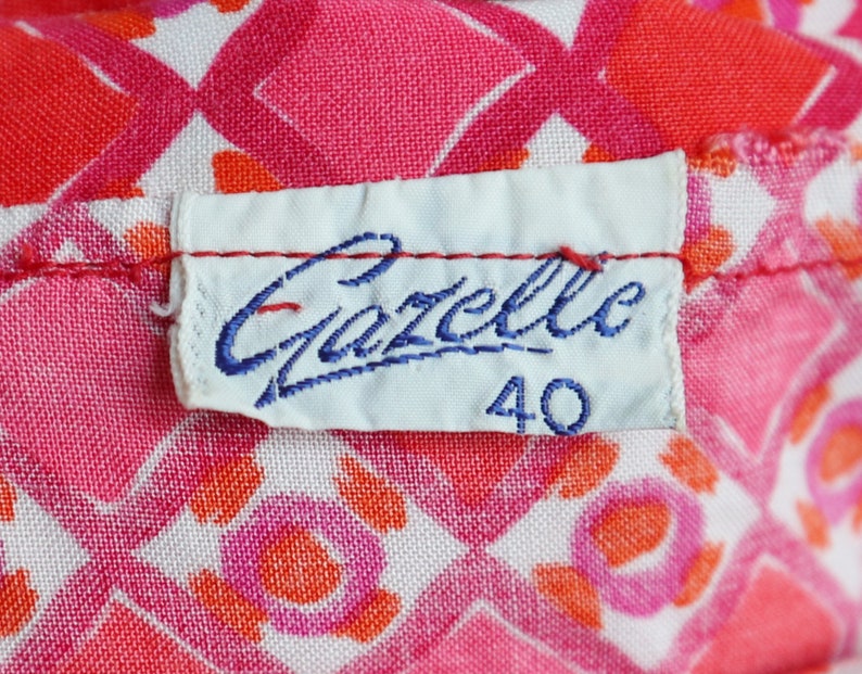 May include: Close-up of a fabric with a repeating pattern of pink, red, and white geometric shapes. A white label with the blue text "Gazelle" and the number "40" is sewn onto the fabric.