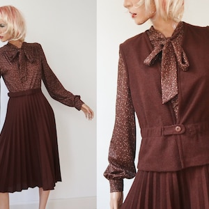 May include: A vintage two-piece outfit featuring a long-sleeved blouse with a bow tie and a pleated skirt. The blouse has a brown floral pattern, and the skirt is a solid dark brown. A matching vest with a button closure completes the ensemble.