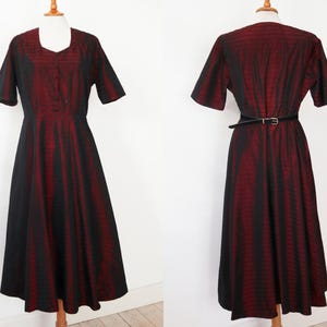 May include: A vintage-style dress with a black and red striped pattern. The dress features short sleeves, a square neckline, and a fitted bodice that flows into a flared skirt. A black belt cinches the waist. The dress is displayed on a mannequin.