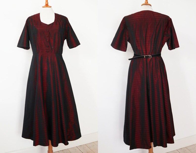 May include: A vintage-style dress with a black and red striped pattern. The dress features short sleeves, a square neckline, and a fitted bodice that flows into a flared skirt. A black belt cinches the waist. The dress is displayed on a mannequin.