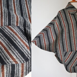 May include: A vintage cape-style jacket with a striped pattern in black, gray, and rust-colored stripes. The jacket features a collar, large buttons, and two front pockets. The fabric has a textured appearance.