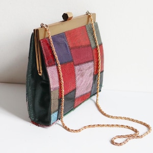 Op de afbeelding: Een vintage patchwork handtas met een goudkleurig frame en kettingriem. De tas heeft een kleurrijk patchwork ontwerp met rode, paarse en zwarte vierkanten. De tas is ongeveer 20 cm breed en 15 cm hoog.