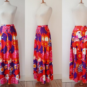 May include: A long, pleated skirt with a vibrant, retro-inspired print. The skirt features a bold pattern of orange, red, purple, and white geometric shapes and floral accents. The skirt is displayed on a mannequin.