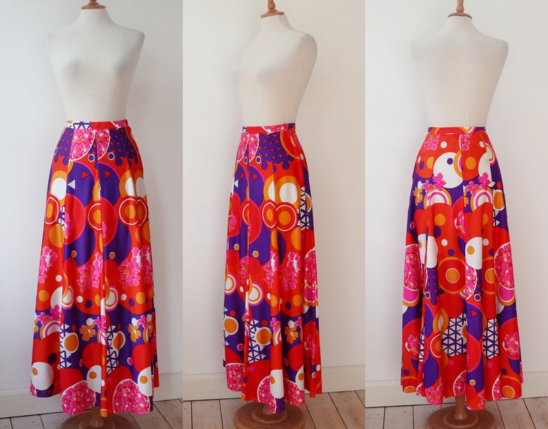 May include: A long, pleated skirt with a vibrant, retro-inspired print. The skirt features a bold pattern of orange, red, purple, and white geometric shapes and floral accents. The skirt is displayed on a mannequin.