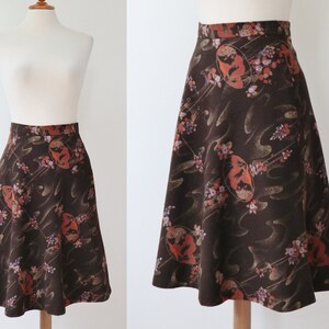 K&ouml;nnte beinhalten: A-Linien-Rock in dunkelbraunem Stoff mit floralem Muster. Der Rock hat eine hohe Taille und eine ausgestellte Silhouette. Das Muster umfasst orange, rosa und wei&szlig;e florale Elemente und abstrakte Formen.