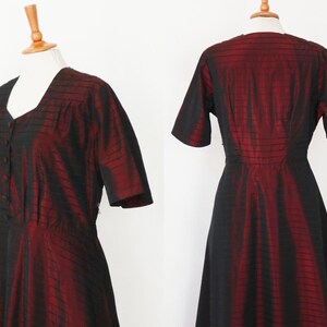 May include: A vintage, knee-length dress in a striking red and black color scheme. The dress features a V-neckline, short sleeves, and a button-down front. The fabric has a subtle sheen, with vertical black stripes over a red base, creating a textured appearance.