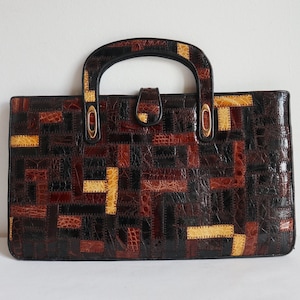 May include: A rectangular handbag with a patchwork design in shades of brown, tan, and black. The bag features a top handle, gold-toned hardware, and a clasp closure. The overall style is vintage.