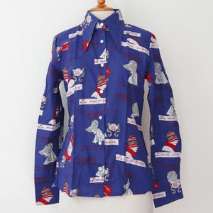 Op de afbeelding: Een blauw overhemd met lange mouwen met een herhalend patroon van illustraties en tekst. De illustraties tonen vrouwen met hoeden en de tekst bevat de woorden "La mode". Het overhemd heeft een puntkraag.