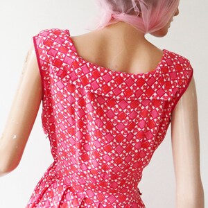 May include: A vintage red and pink patterned dress. The dress features a geometric design of squares and hearts. The dress has a scoop neckline and a fitted bodice. The dress is sleeveless and appears to be made of a lightweight fabric.