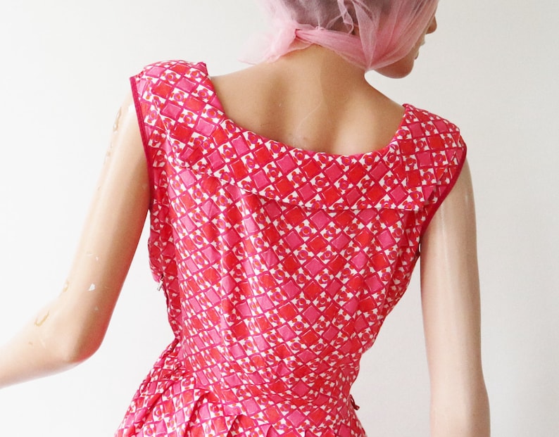May include: A vintage red and pink patterned dress. The dress features a geometric design of squares and hearts. The dress has a scoop neckline and a fitted bodice. The dress is sleeveless and appears to be made of a lightweight fabric.