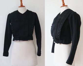 May include: Black cropped bolero jacket with long sleeves. The jacket features a V-neck, decorative trim, and small buttons down the front. The sleeves and front panels have intricate embroidered detailing. The jacket is displayed on a mannequin.