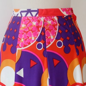 May include: A vibrant, high-waisted skirt featuring a bold geometric pattern. The design incorporates orange, purple, pink, and white shapes, including circles, triangles, and abstract floral elements. The skirt has a pleated front and a bright red waistband.