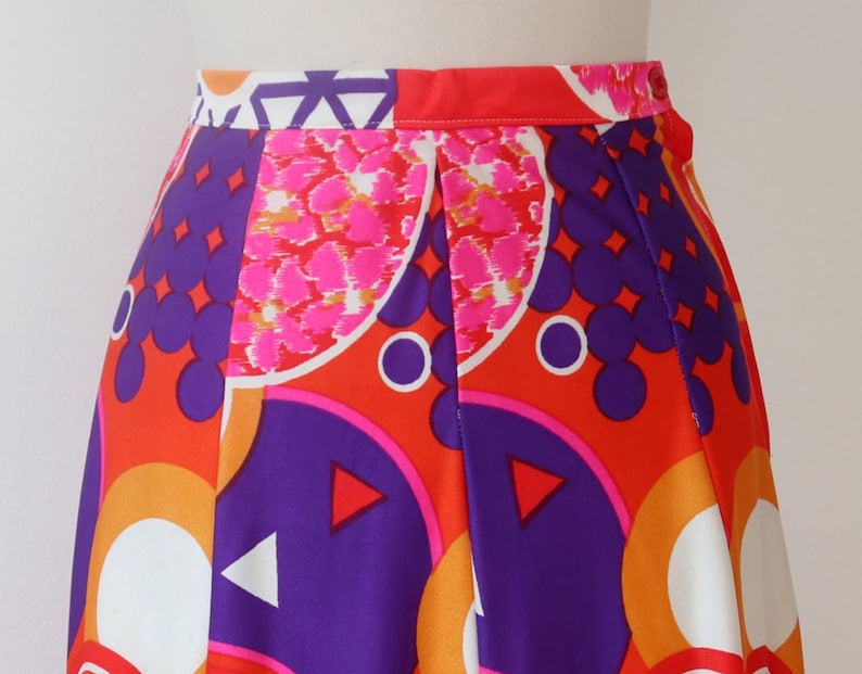 May include: A vibrant, high-waisted skirt featuring a bold geometric pattern. The design incorporates orange, purple, pink, and white shapes, including circles, triangles, and abstract floral elements. The skirt has a pleated front and a bright red waistband.
