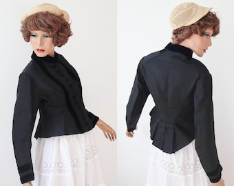 May include: Black jacket with velvet trim and buttons. The jacket has long sleeves and a high collar. The model is wearing a straw hat and a white skirt. The jacket is a vintage style.
