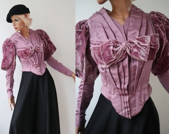 May include: A vintage-style mauve velvet top with a large bow detail and pleated bodice. The top features long sleeves and dramatic, puffed sleeves. Paired with a long black skirt, the ensemble is displayed on a mannequin.