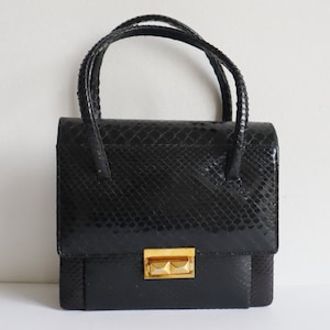 Puede incluir: Un bolso de mano negro con un patrón de piel de serpiente texturizado. El bolso tiene forma rectangular con cierre de solapa y un cierre dorado. Cuenta con dos asas pequeñas en la parte superior.