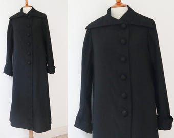 Può includere: Un cappotto lungo nero con un ampio colletto e grandi bottoni rotondi. Il cappotto è esposto su un manichino e ha maniche lunghe con polsini ripiegati. Il cappotto sembra essere realizzato in un tessuto liscio, forse lana.