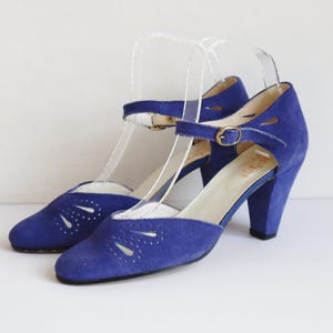 Könnte beinhalten: Ein Paar blaue Vintage-Wildleder-Heels mit Riemen und Schnallenverschluss. Die Schuhe haben ein Peep-Toe-Design mit dekorativen Aussparungen und einem klobigen Absatz. Die Absätze sind etwa 7,6 cm hoch.