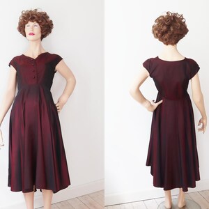 May include: A vintage burgundy dress with a flared skirt and cap sleeves. The dress features a button-up bodice and a subtle ombre effect, transitioning from a darker shade at the top to a lighter hue towards the hem. The dress is displayed on a mannequin.