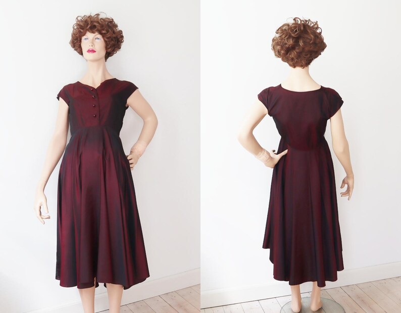 May include: A vintage burgundy dress with a flared skirt and cap sleeves. The dress features a button-up bodice and a subtle ombre effect, transitioning from a darker shade at the top to a lighter hue towards the hem. The dress is displayed on a mannequin.