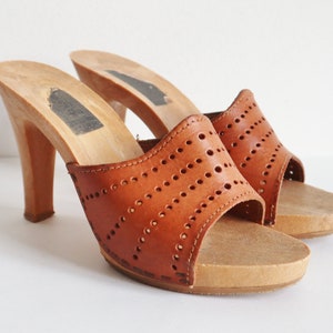 May include: A pair of vintage brown leather clogs with wooden heels. The open-toe sandals feature a perforated design on the upper and a platform sole. The heels are approximately 7.6 cm tall.
