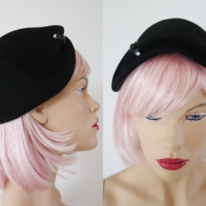 May include: A black hat with a curved design, adorned with a small, decorative jewel. The hat is made of a smooth material and is shown from multiple angles. The hat is a vintage style.