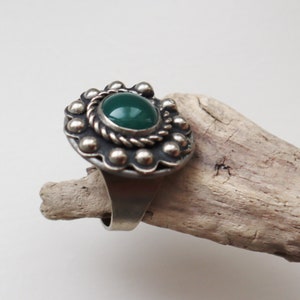 May include: A silver ring featuring a large, oval, dark green stone. The stone is encircled by a twisted rope design and a border of small silver beads. The ring is displayed on a piece of driftwood.