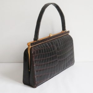以下が含まれることがあります： クロコダイルのような質感のヴィンテージブラウンハンドバッグ。長方形のバッグは、ゴールドトーンの留め金と、お揃いのハンドルが特徴です。バッグの長さは約25cm、高さは約15cmです。