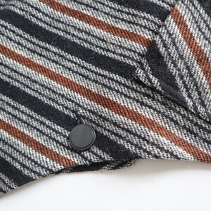 May include: Close-up of a section of a black, white, and brown striped fabric. The fabric has a diagonal pattern with black, white, and rust-colored stripes. A black button is visible on the edge of the fabric, suggesting it's part of a garment.