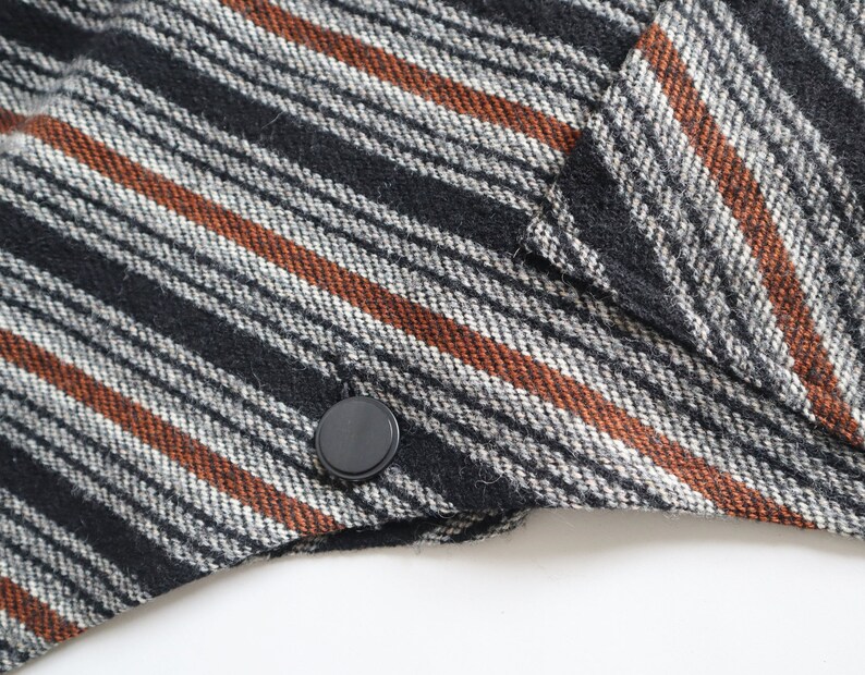 May include: Close-up of a section of a black, white, and brown striped fabric. The fabric has a diagonal pattern with black, white, and rust-colored stripes. A black button is visible on the edge of the fabric, suggesting it's part of a garment.