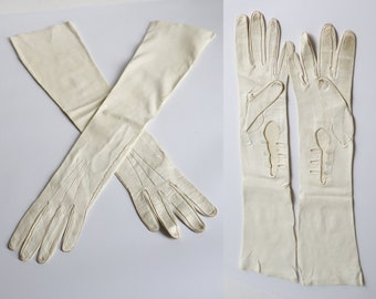 May include: A pair of off-white, long opera gloves. The gloves are made of a soft material and feature decorative cutouts on the back of the hands. The gloves are long, extending well past the elbows.