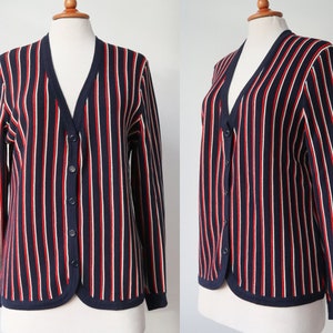 Op de afbeelding: Marineblau vest met verticale rode en witte strepen. De trui met lange mouwen heeft een V-hals, een knoopsluiting en een marineblauwe bies. Het kledingstuk wordt op een mannequin gepresenteerd.
