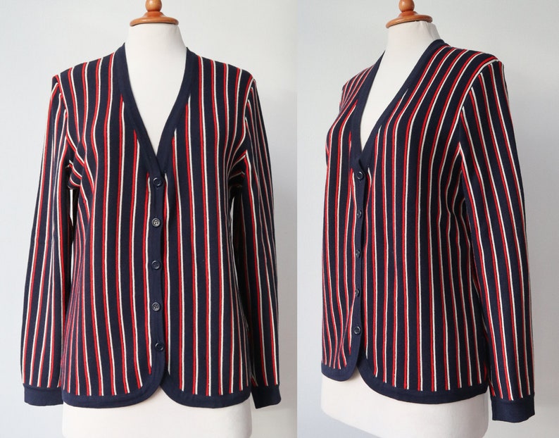 Op de afbeelding: Marineblau vest met verticale rode en witte strepen. De trui met lange mouwen heeft een V-hals, een knoopsluiting en een marineblauwe bies. Het kledingstuk wordt op een mannequin gepresenteerd.