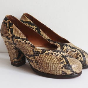 Può includere: Un paio di décolleté vintage con motivo a pelle di serpente. Le scarpe presentano una palette di colori neutri di beige, marrone e nero, con un aspetto strutturato. I tacchi sono spessi e squadrati e le scarpe hanno una punta arrotondata.