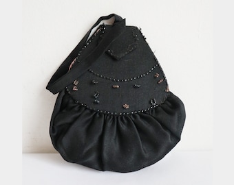 May include: A black vintage handbag with a rounded bottom and a small flap. The bag features a gathered skirt-like detail and is embellished with small beads. It has a black strap for carrying.