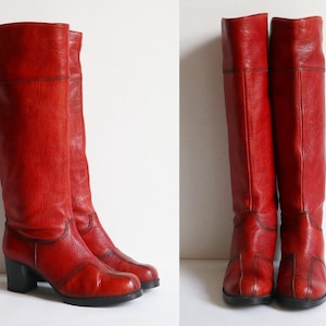 May include: A pair of knee-high red leather boots featuring a textured finish, a rounded toe, and a block heel. The boots have visible stitched seam detailing along the foot and shaft, with a solid black heel and sole construction.
