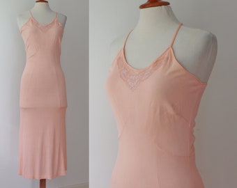 May include: A peach-colored slip dress with a halter neckline and delicate lace detailing at the bust. The dress is displayed on a mannequin, showcasing its form-fitting silhouette and midi length. The fabric appears soft and lightweight.