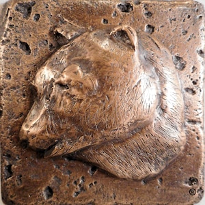 May include: A bronze sculpture of a bear's head in profile, with a textured surface that resembles fur. The sculpture is mounted on a square, textured base.