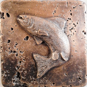 Puede incluir: Un azulejo de color bronce con una imagen en relieve de un pez saltando fuera del agua. El azulejo tiene una superficie rugosa y texturizada.