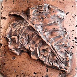 May include: A copper-coloured metal sculpture of a leaf with a textured surface. The sculpture is set into a square, copper-coloured tile.