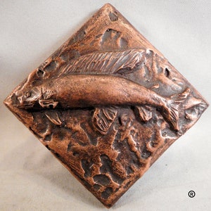 Puede incluir: Un azulejo de color cobre con un diseño en relieve texturizado de un pez nadando. El azulejo tiene una superficie rugosa y texturizada y una forma de diamante.