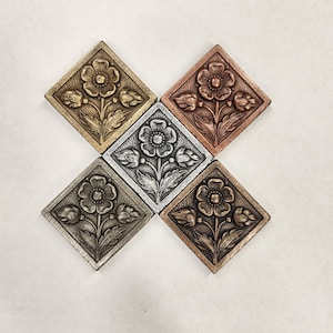 Może przedstawiać: Widok z góry na pięć metalowych płytek w kształcie rombu, z których każda ma kwiatowy wzór. Płytki ułożone są w kształt krzyża, z centralną płytką w kolorze srebrnym, a otaczającymi w odcieniach złota, brązu i miedzi.