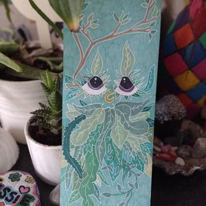 Puede incluir: Un marcapáginas pintado a mano con una criatura verde caprichosa con ojos grandes y una barba de hojas. La criatura está rodeada de una variedad de hojas y enredaderas verdes sobre un fondo azul claro.