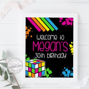 May include: A framed black sign with the text "Welcome to Megan's 30th birthday" in pink and white. The sign features a colorful Rubik's Cube graphic and neon paint splatters. The top of the sign has a rainbow stripe design.